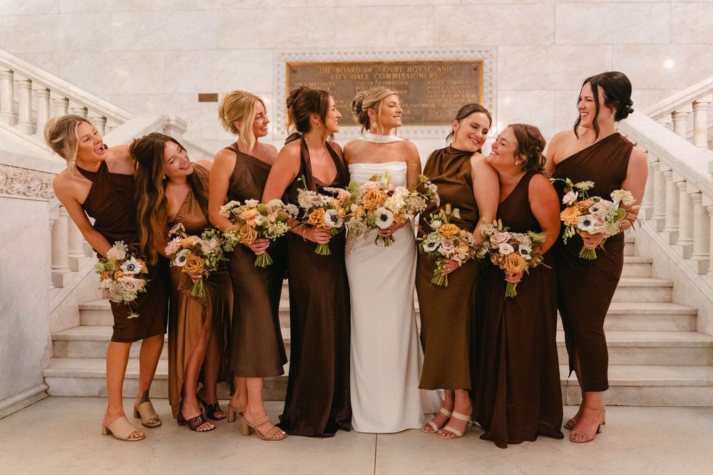 Wedding party in elegant brown and white dresses pose with floral bouquets on grand marble staircase.
