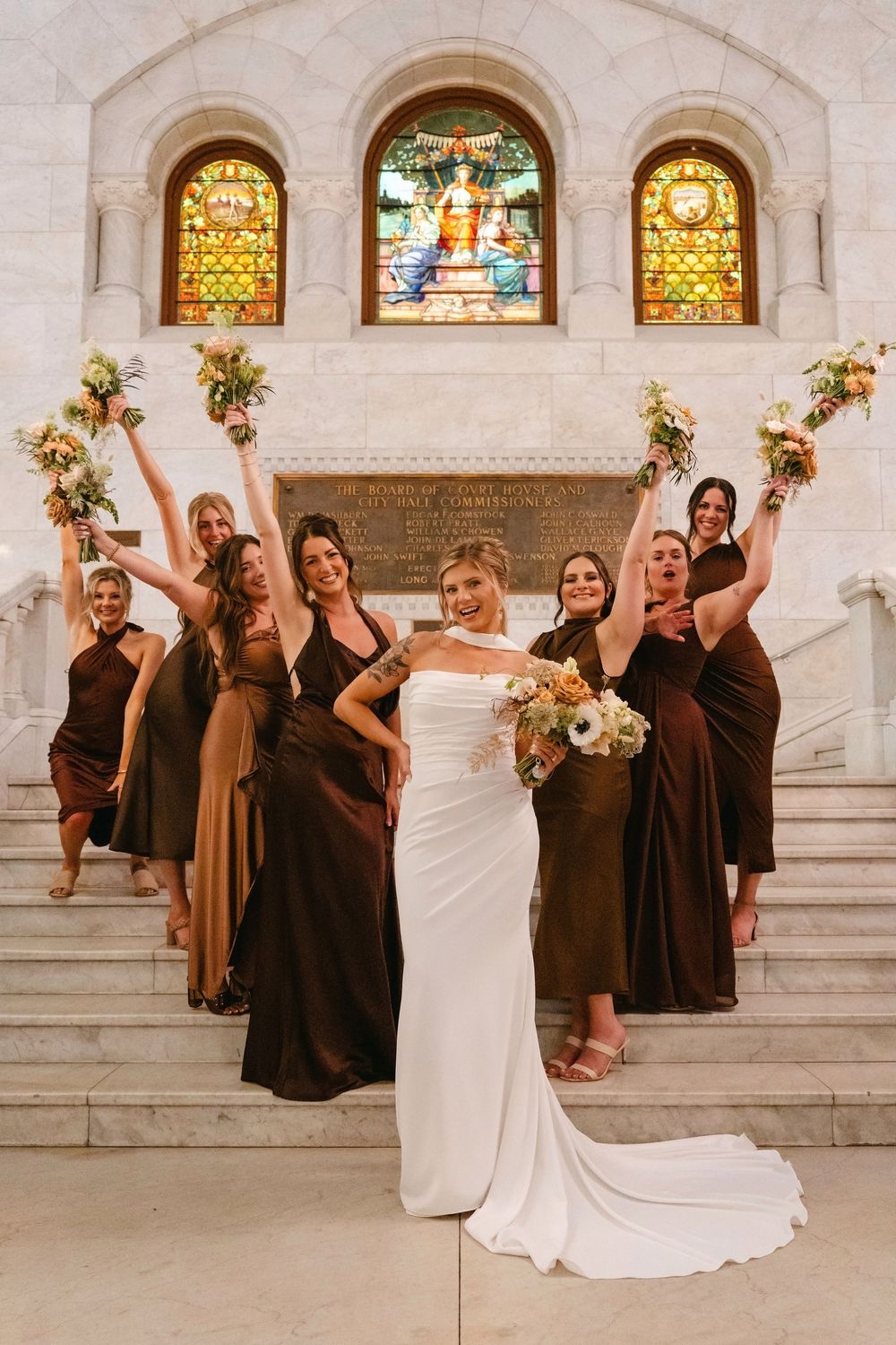 Bridal party in brown dresses strikes fun poses on grand marble staircase with stained glass windows.