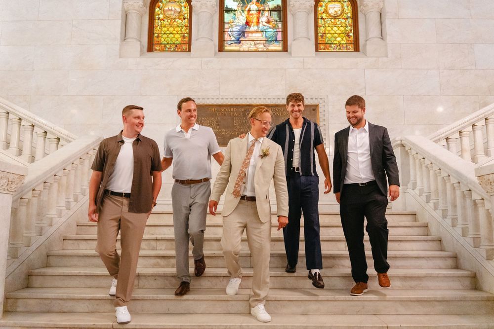Wedding party members walk down an elegant white marble staircase in a grand venue with stained glass windows.