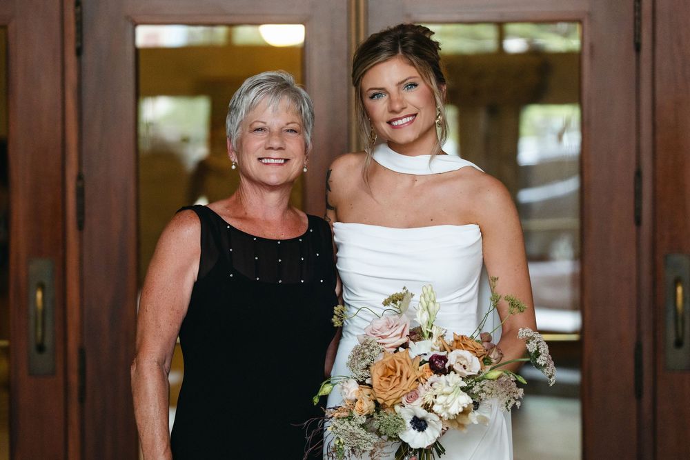 Two women pose together at a formal event, one wearing a black dress and one in a white wedding gown holding a bouquet.