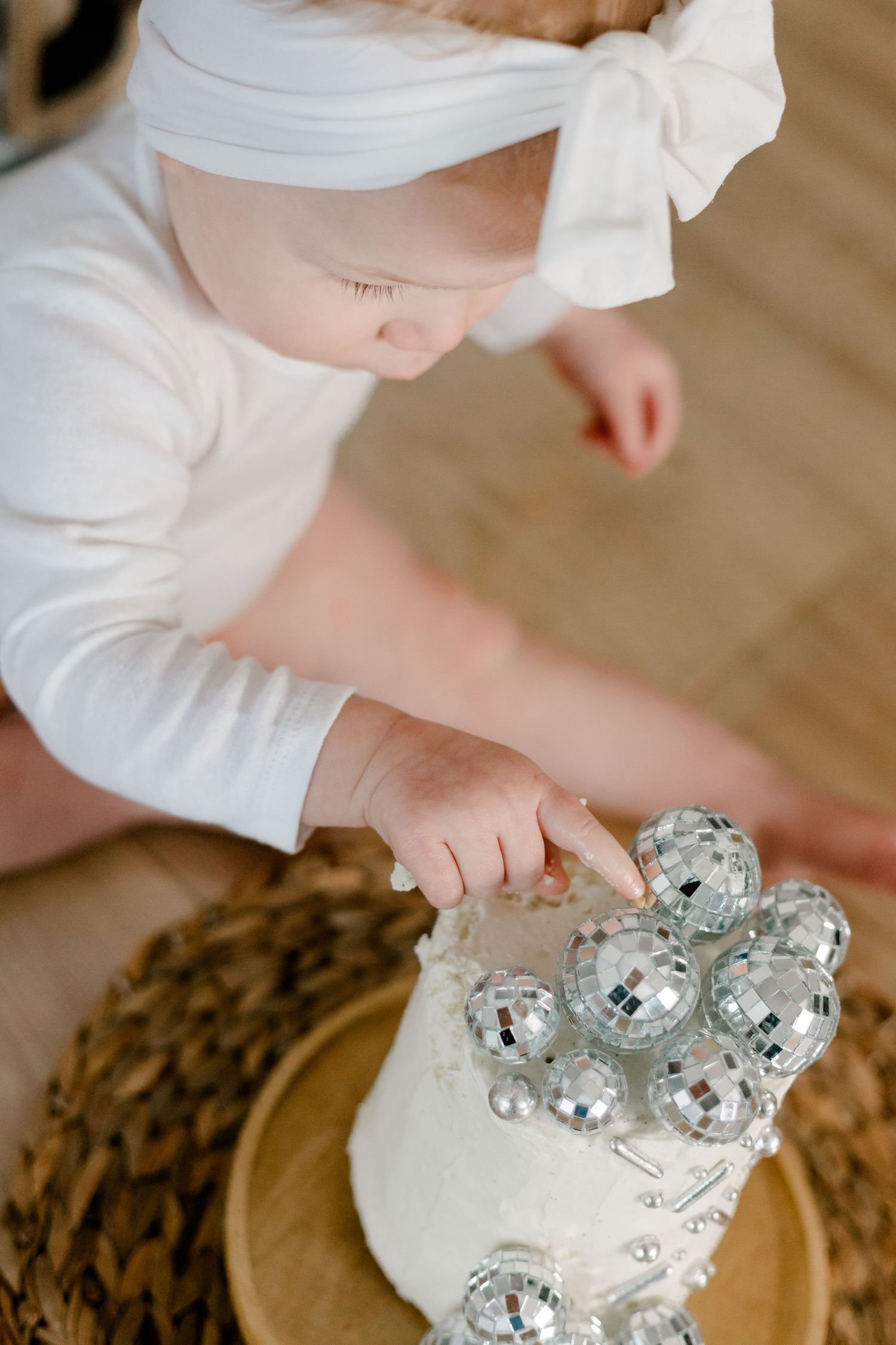 Baby girl in white outfit with bow headband touching disco ball decorated smash cake.