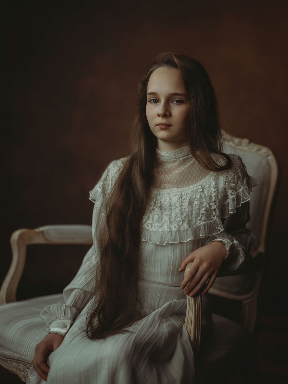 A vintage-style portrait of someone in a Victorian white lace dress with long brown hair sitting in an antique chair.