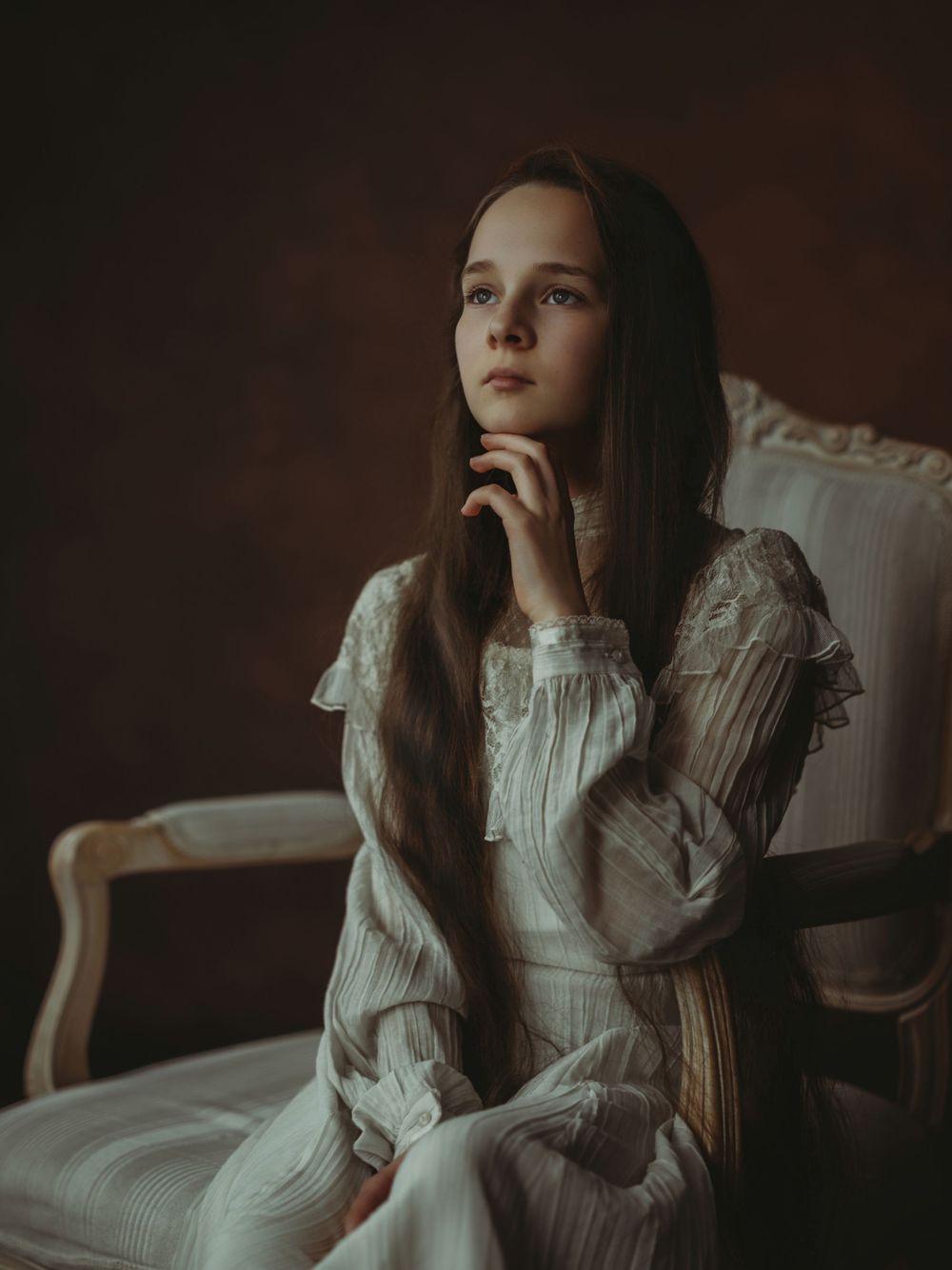 A moody portrait of someone in a vintage white lace dress sitting thoughtfully in an antique chair against a dark background.