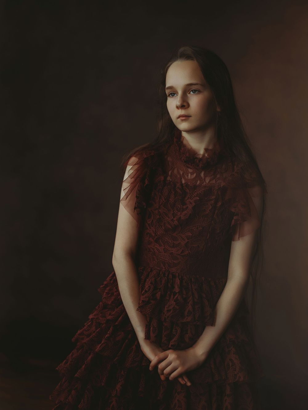 Moody portrait photograph of figure in dark burgundy lace dress against shadowy background in vintage style.