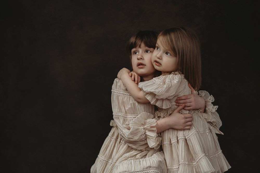 Two young children in vintage white dresses share an emotional embrace against a dark background.