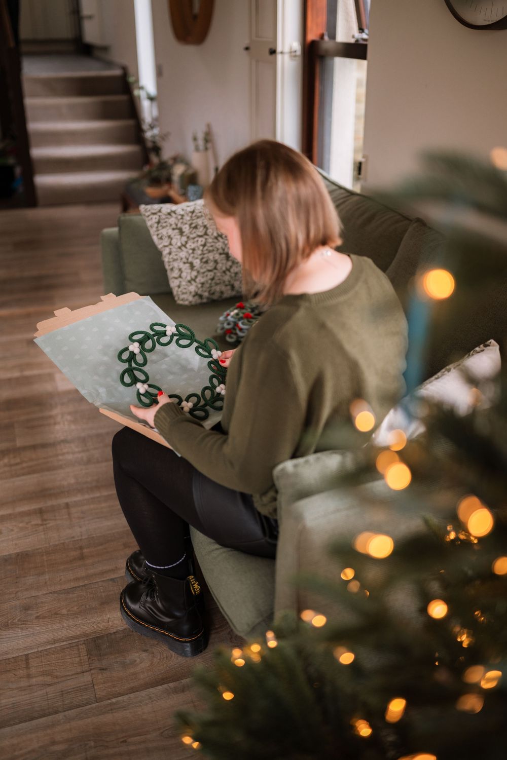 Person in olive green sweater sitting near a decorated Christmas tree with bokeh lights.