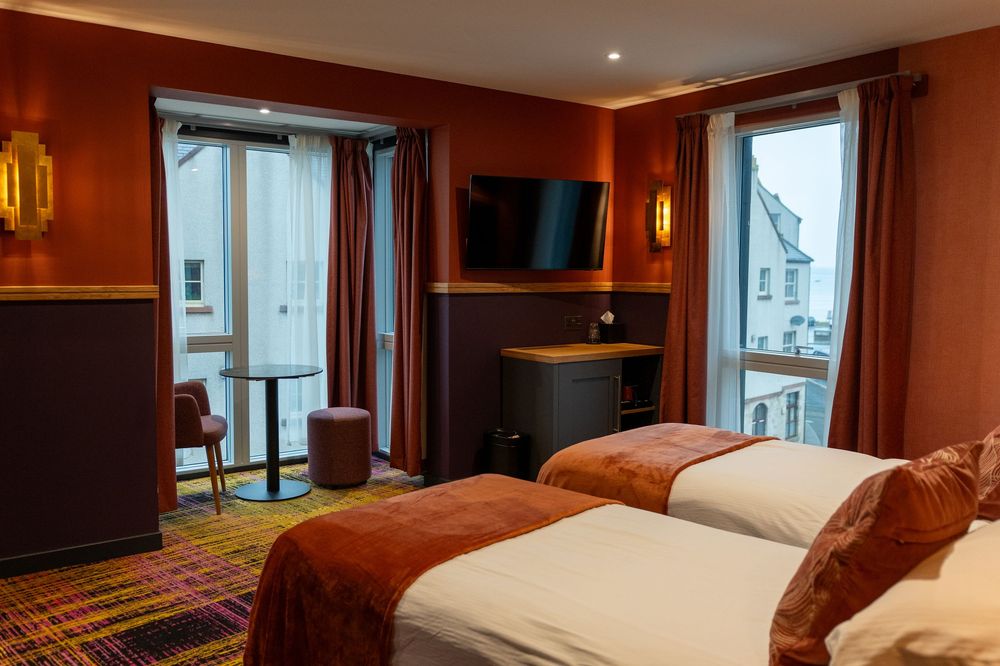 Modern hotel room interior with warm wood paneling, white bedding, and large windows offering city views.