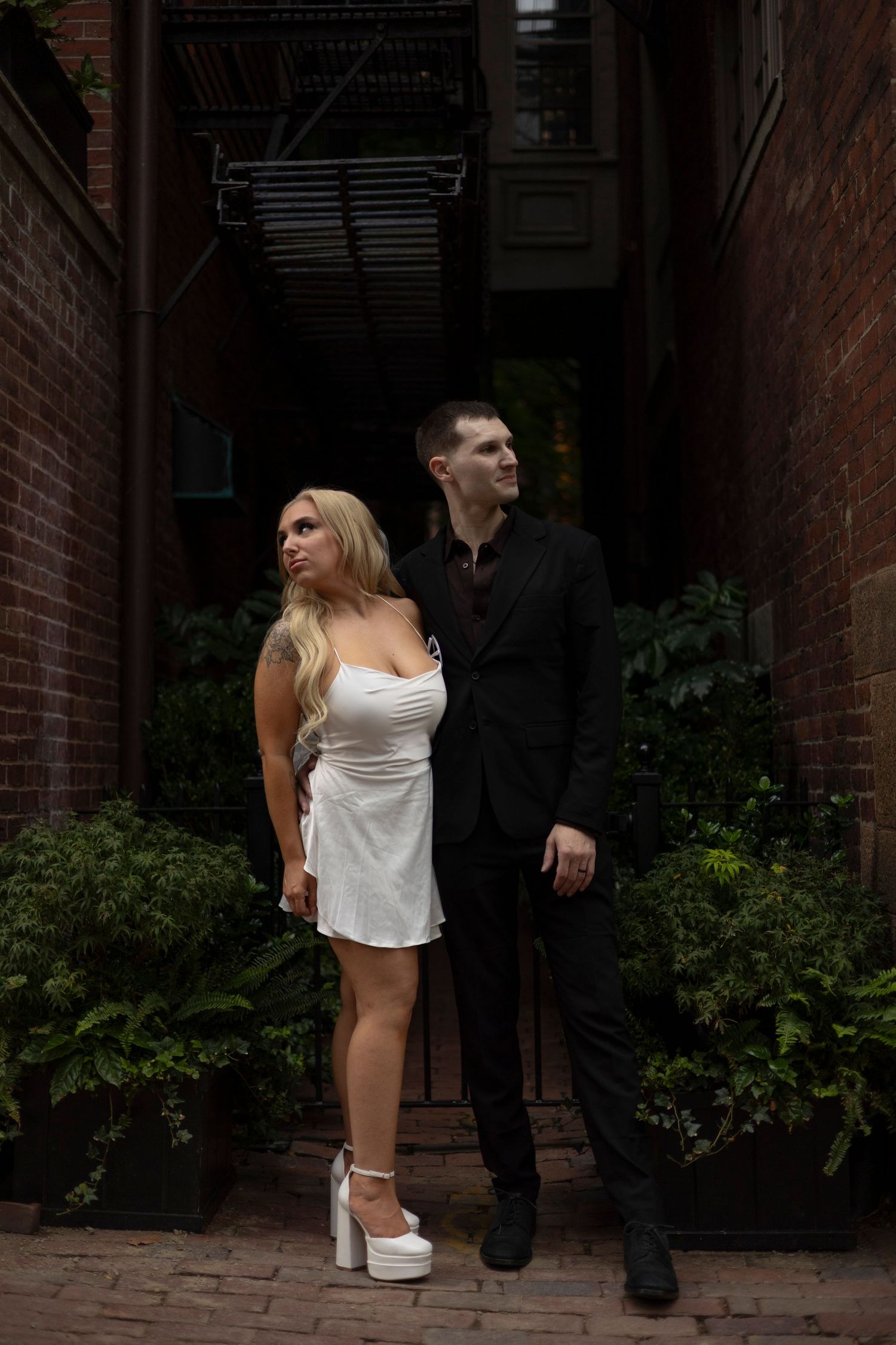A person in a white top and black pants stands in front of a brick building entrance.