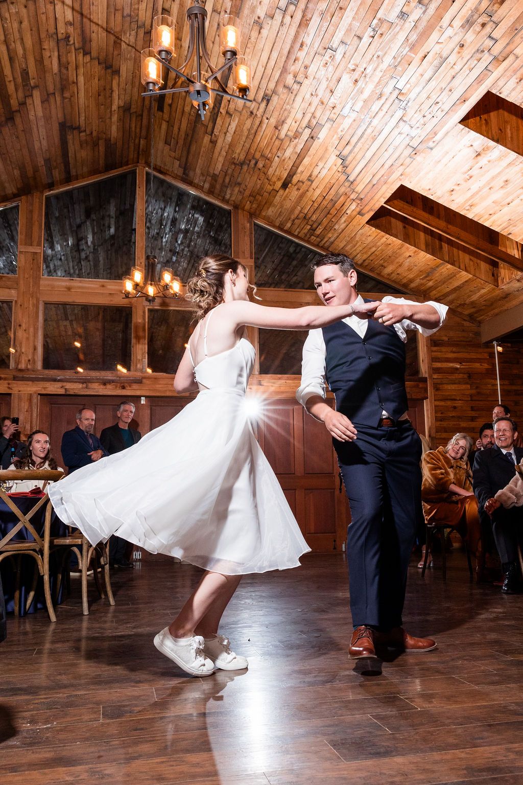 A couple performs an elegant dance spin on a rustic wooden dance floor beneath a vaulted barn ceiling with warm lighting.