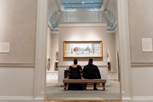 Two people sit on a bench viewing artwork in a white-walled gallery space with hardwood floors.
