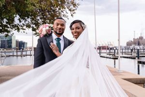 A couple shares a romantic moment on a waterfront boardwalk with boats and a marina in the background.
