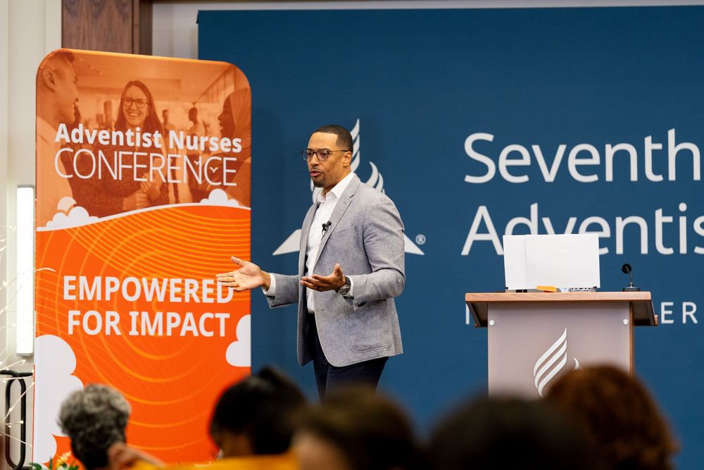 A speaker presents at a conference with orange and blue banners visible in the background.