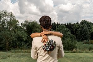 A couple embraces while looking out at a scenic green field and forest landscape on a cloudy day.