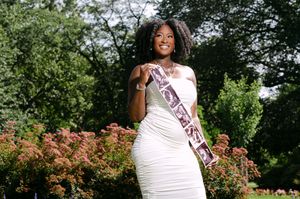A person in a white formal dress poses outdoors in a garden setting with blooming flowers and greenery.