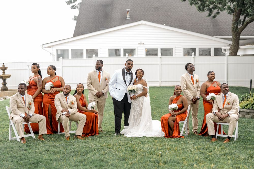 Wedding party in orange and cream colored attire poses together on a grassy lawn for outdoor photos.