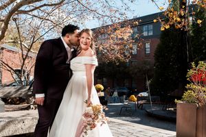 Newlyweds share romantic moments in an autumn courtyard setting with brick buildings and fall foliage in the background.