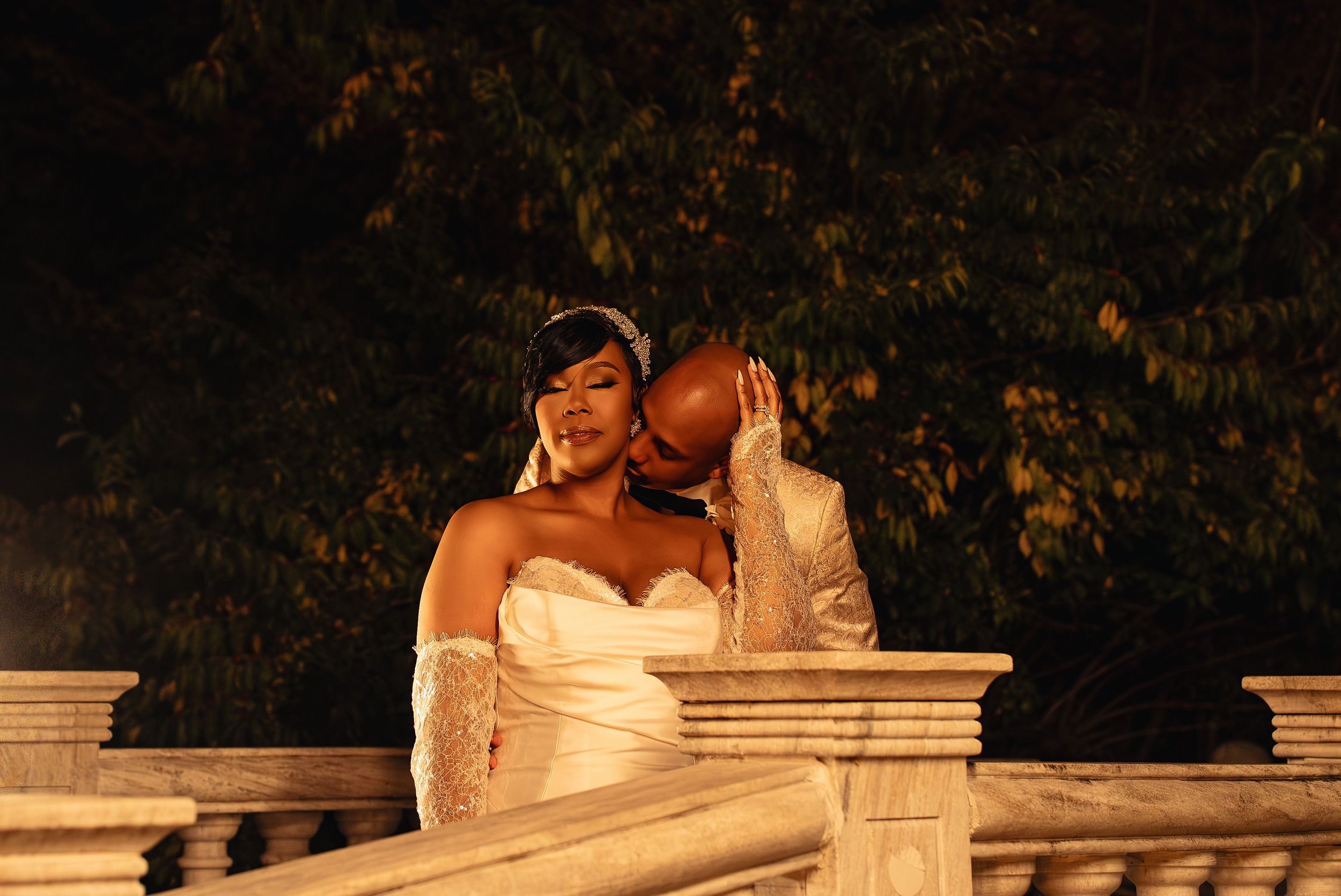 A romantic wedding couple embraces and kisses on a stone balustrade at sunset during their outdoor photoshoot.