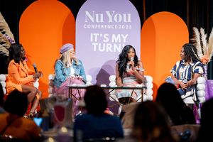 Panel discussion on stage with four speakers seated in front of orange arches and purple NuYou conference backdrop.