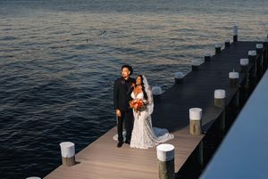 A couple in wedding attire stands on a wooden dock overlooking a serene lake at sunset.