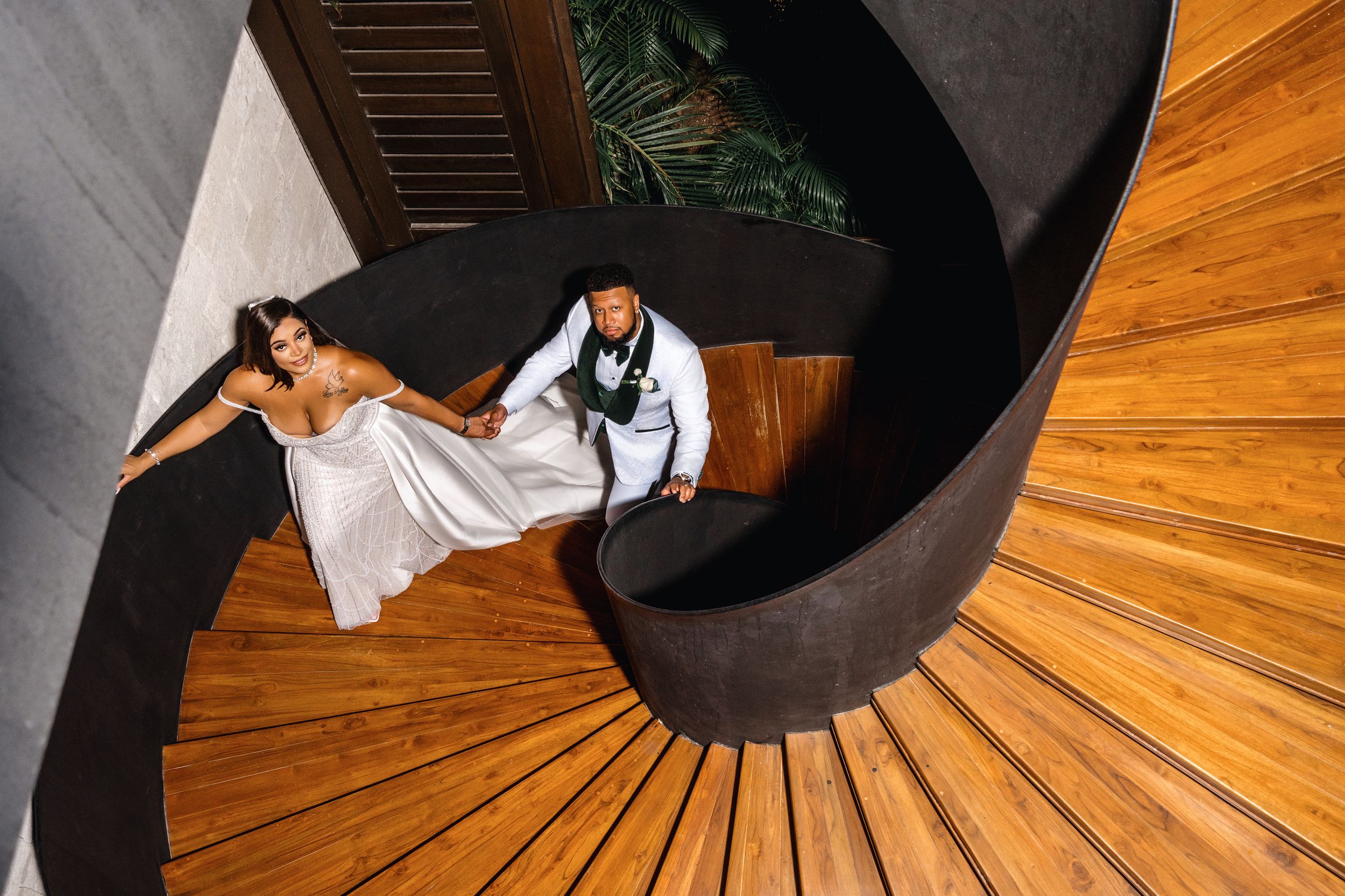 A bride and groom pose on a dramatic spiral staircase with wooden steps and dark curved walls.