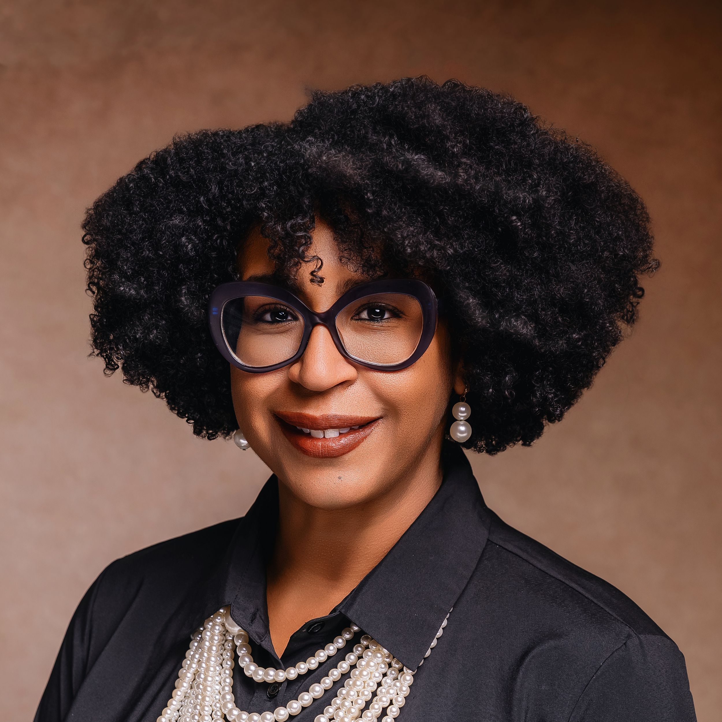 Professional studio portrait with black blazer, pearl necklaces and natural curly hair against brown background.