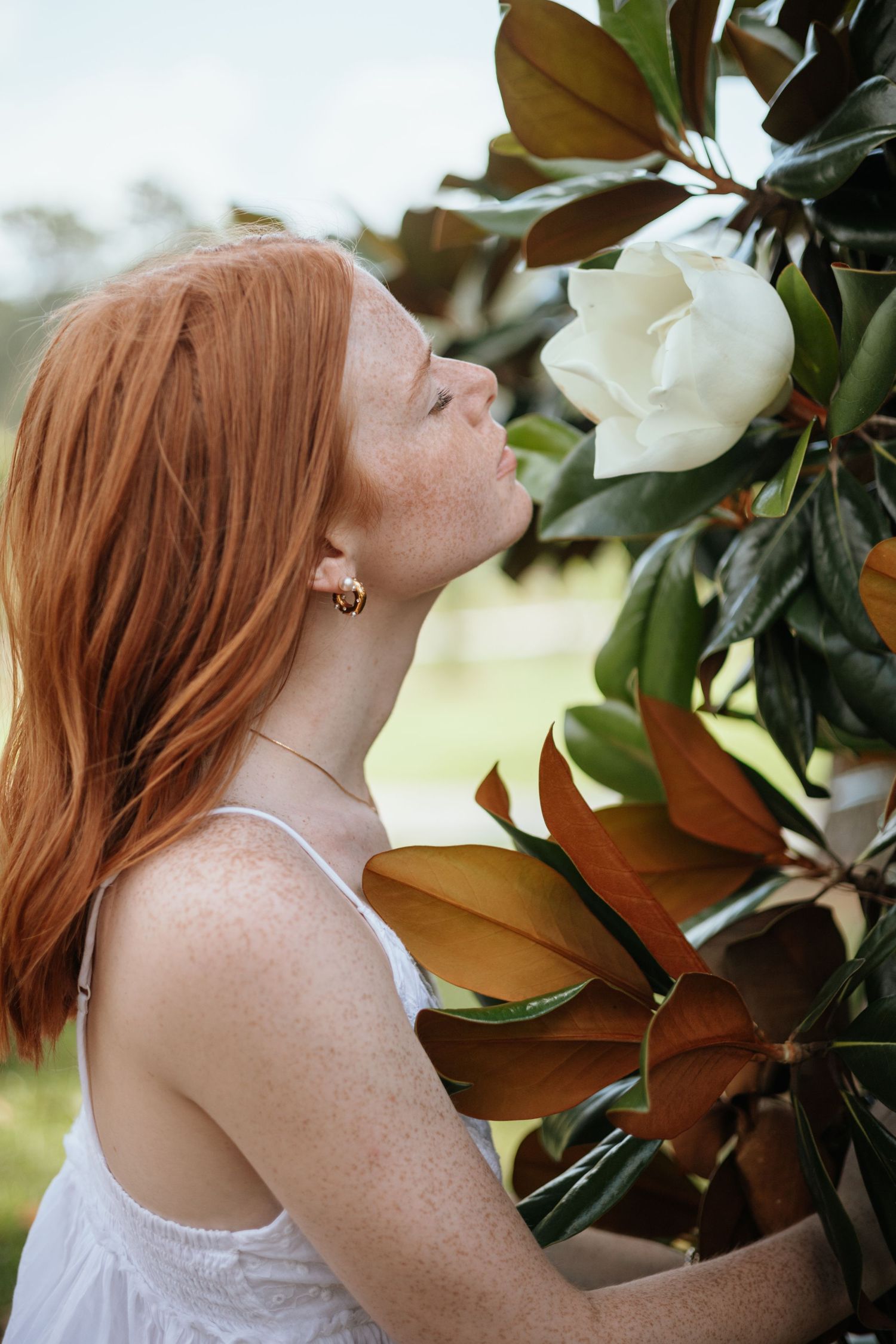 Woman with auburn hair poses near magnolia leaves in white lace top.