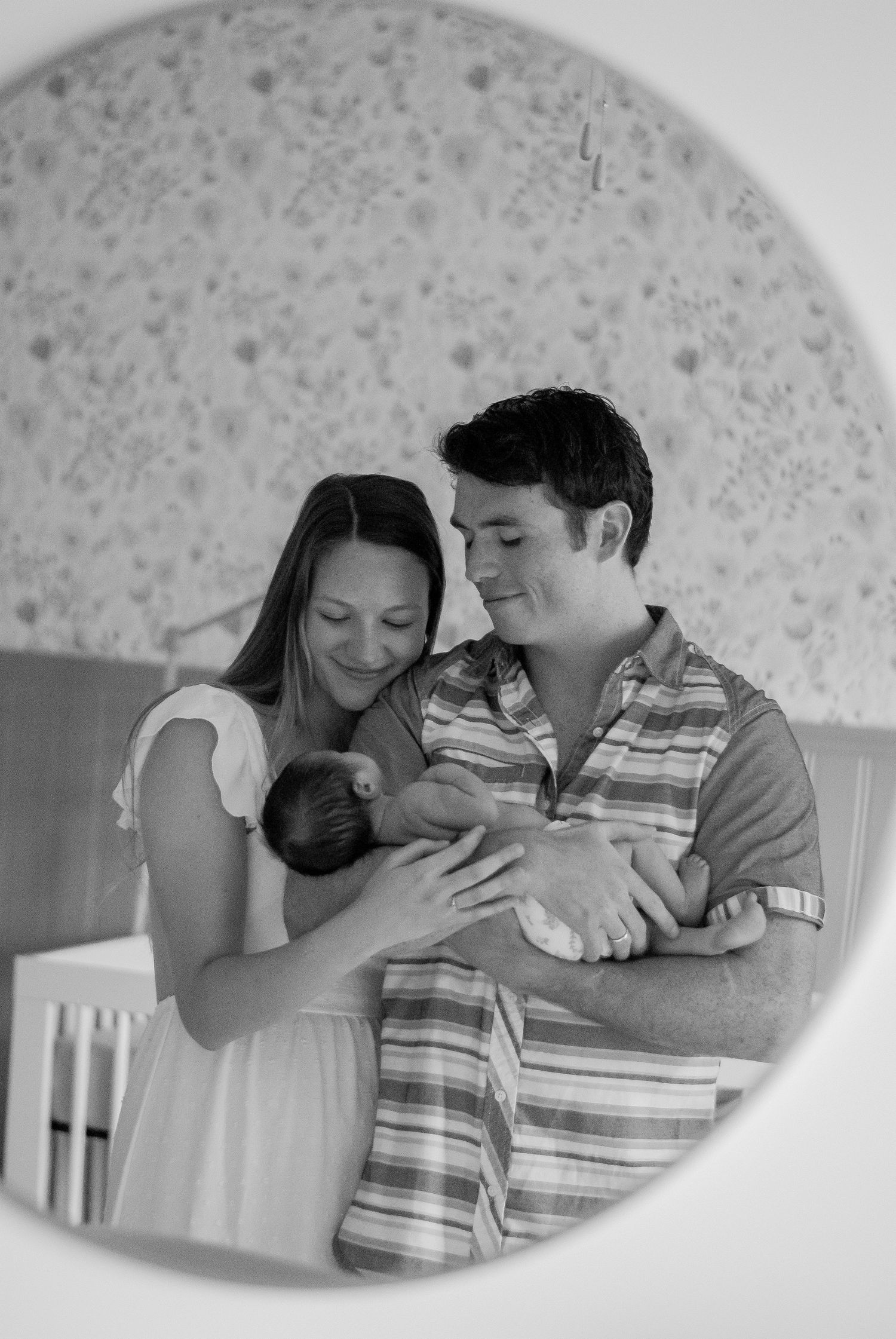A black and white photo showing a couple lovingly embracing their newborn in an intimate home setting reflected in a mirror in the nursery