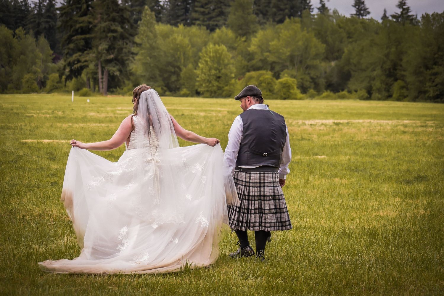 A couple walks together across a scenic meadow with evergreen trees in the background during a summer day.
