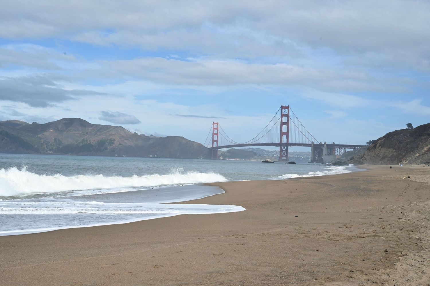 Panoramic view of Baker Beach with the Golden Gate Bridge visible in the distance on a cloudy day in San Francisco, California.