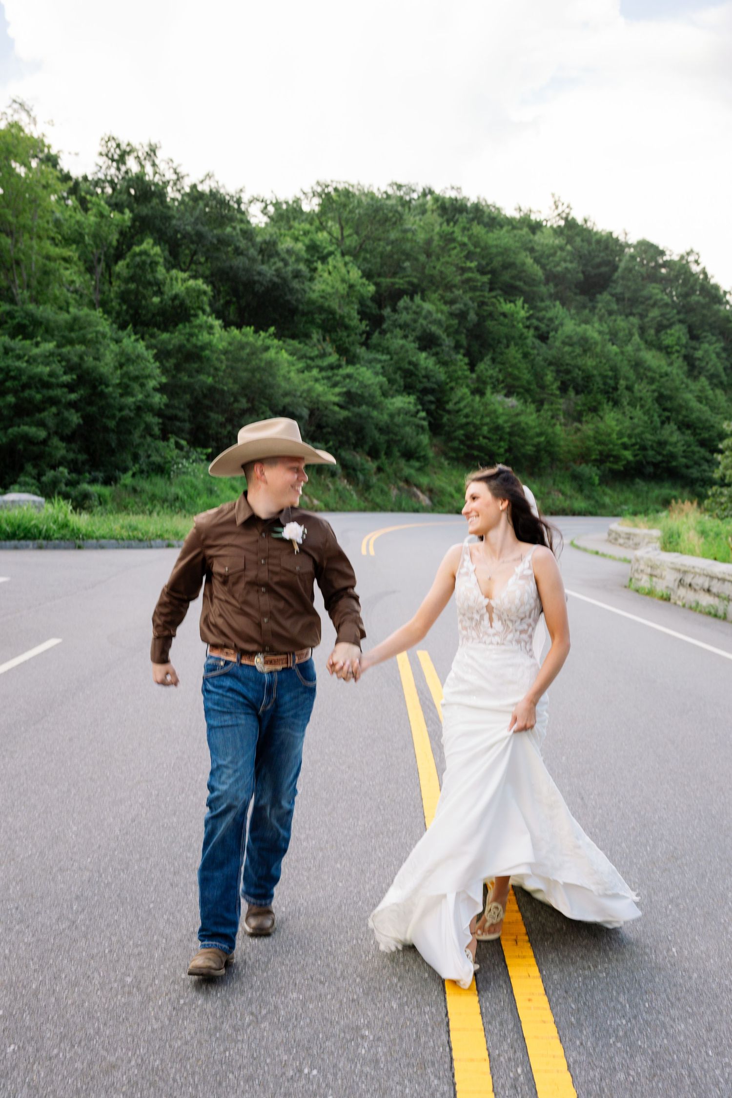 A couple playfully walks and dances along a double yellow line on an empty rural road surrounded by green trees.