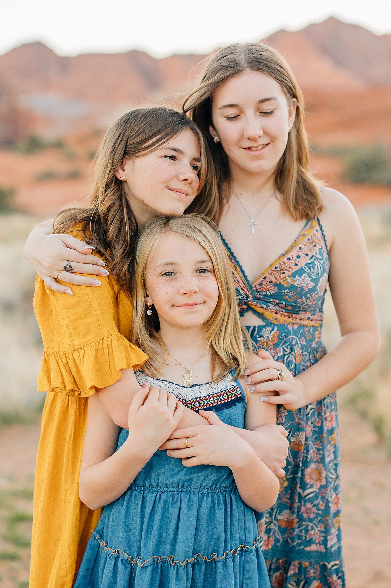Two people wearing blue and yellow dresses embrace while standing in front of red desert rock formations at sunset.
