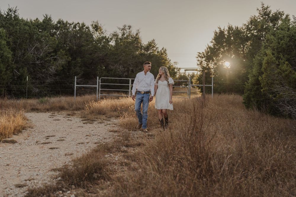 Couple walking hand in hand through a rustic outdoor field at golden hour with sun flare in the background.