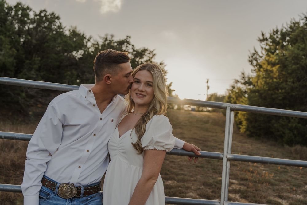 A romantic couple embraces by a rustic wooden fence at sunset in a rural countryside setting.