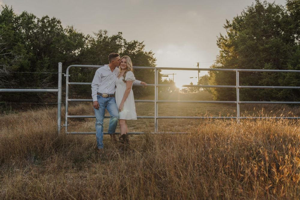 Romantic sunset engagement session with couple sitting on rustic metal gate in golden field.