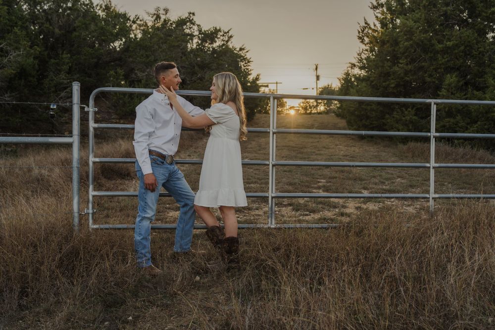 A couple shares a romantic moment at sunset by a rustic wooden fence in a rural countryside setting.