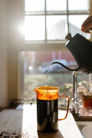Coffee being poured into a clear glass mug creating a golden cascade effect near a bright window.