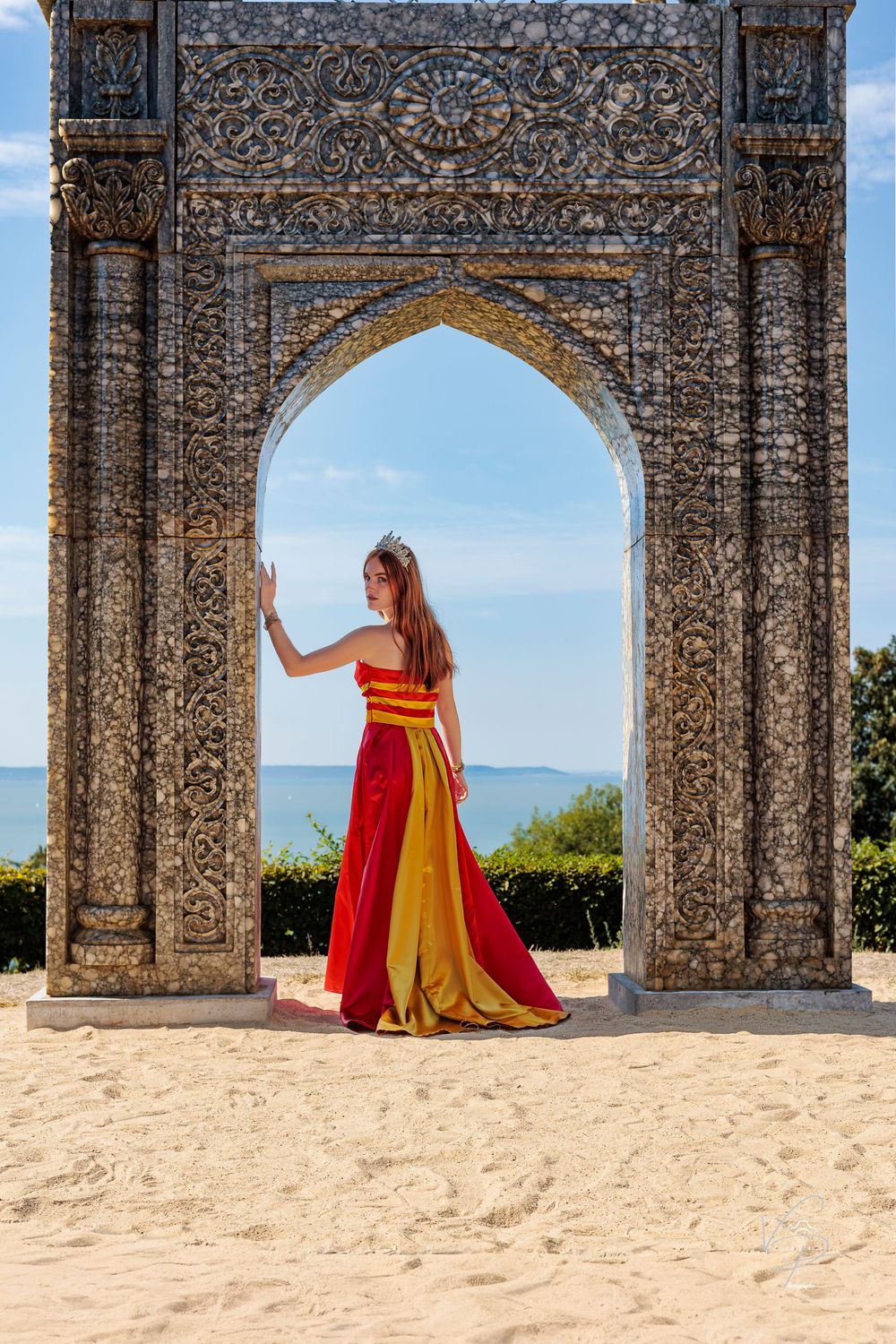 Dramatic pose in red and gold dress framed by ornate stone architecture.