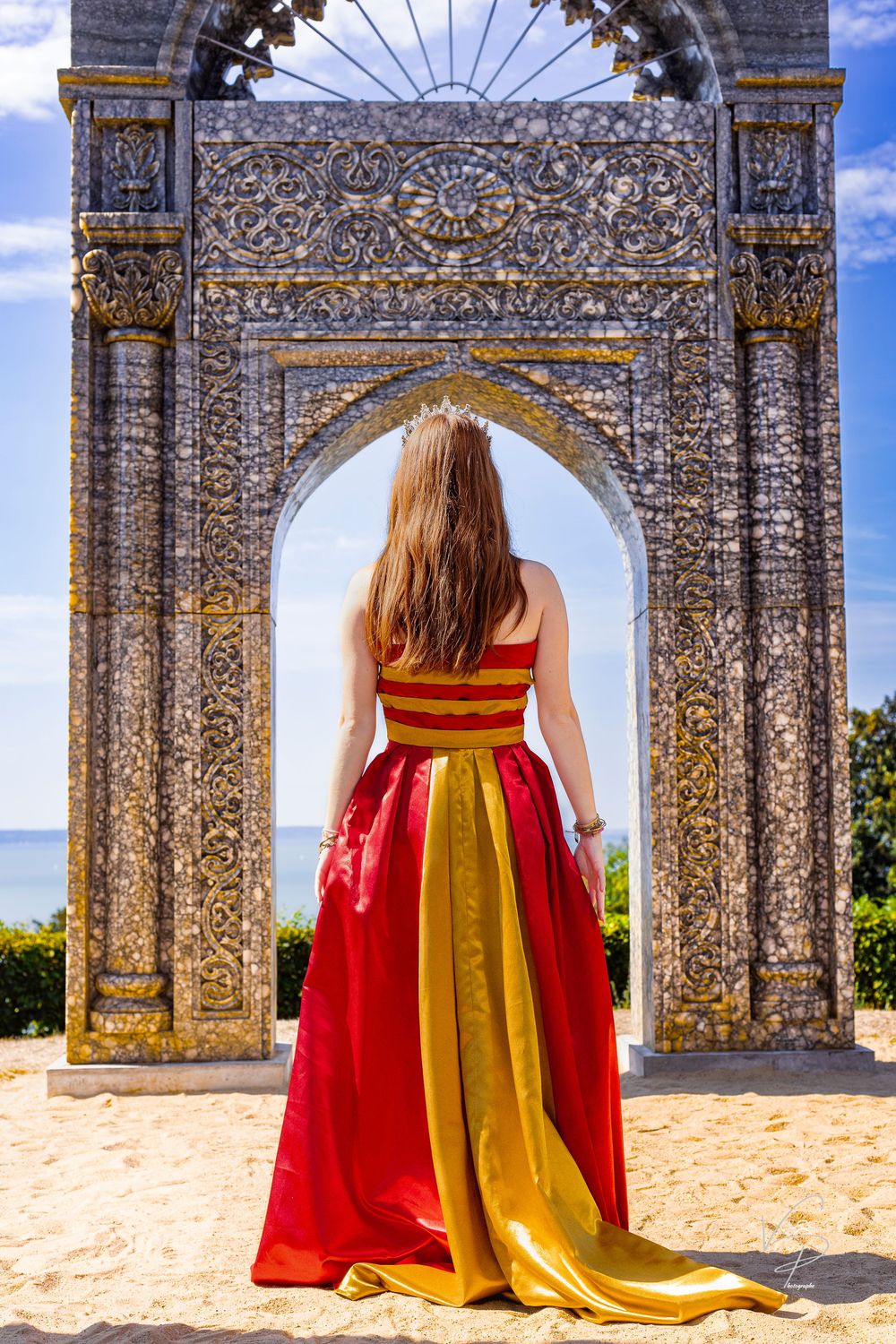 Red and gold dress flows majestically through stone archways against blue sky.