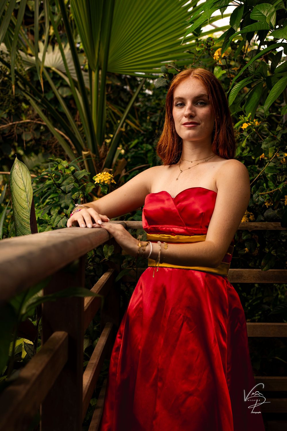 Model in red dress poses elegantly against tropical foliage backdrop in garden setting.