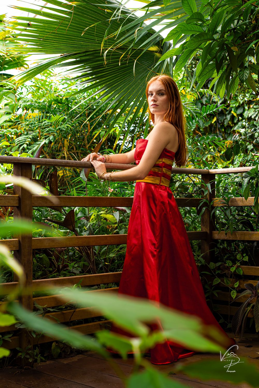 Woman in flowing red dress reclines on wooden railing surrounded by lush palm fronds.
