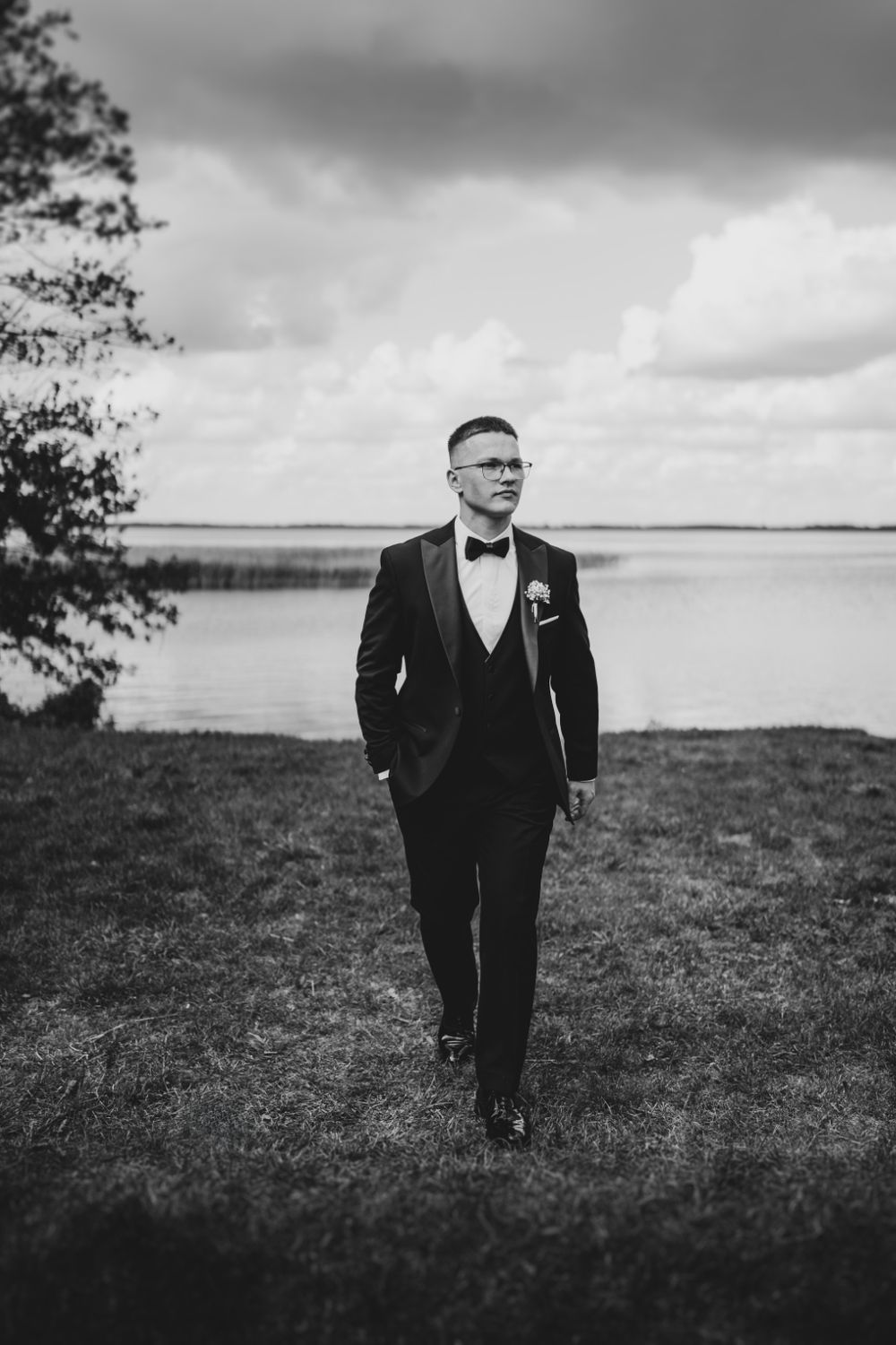 Groom in black tuxedo walks along waterfront under dramatic sky.