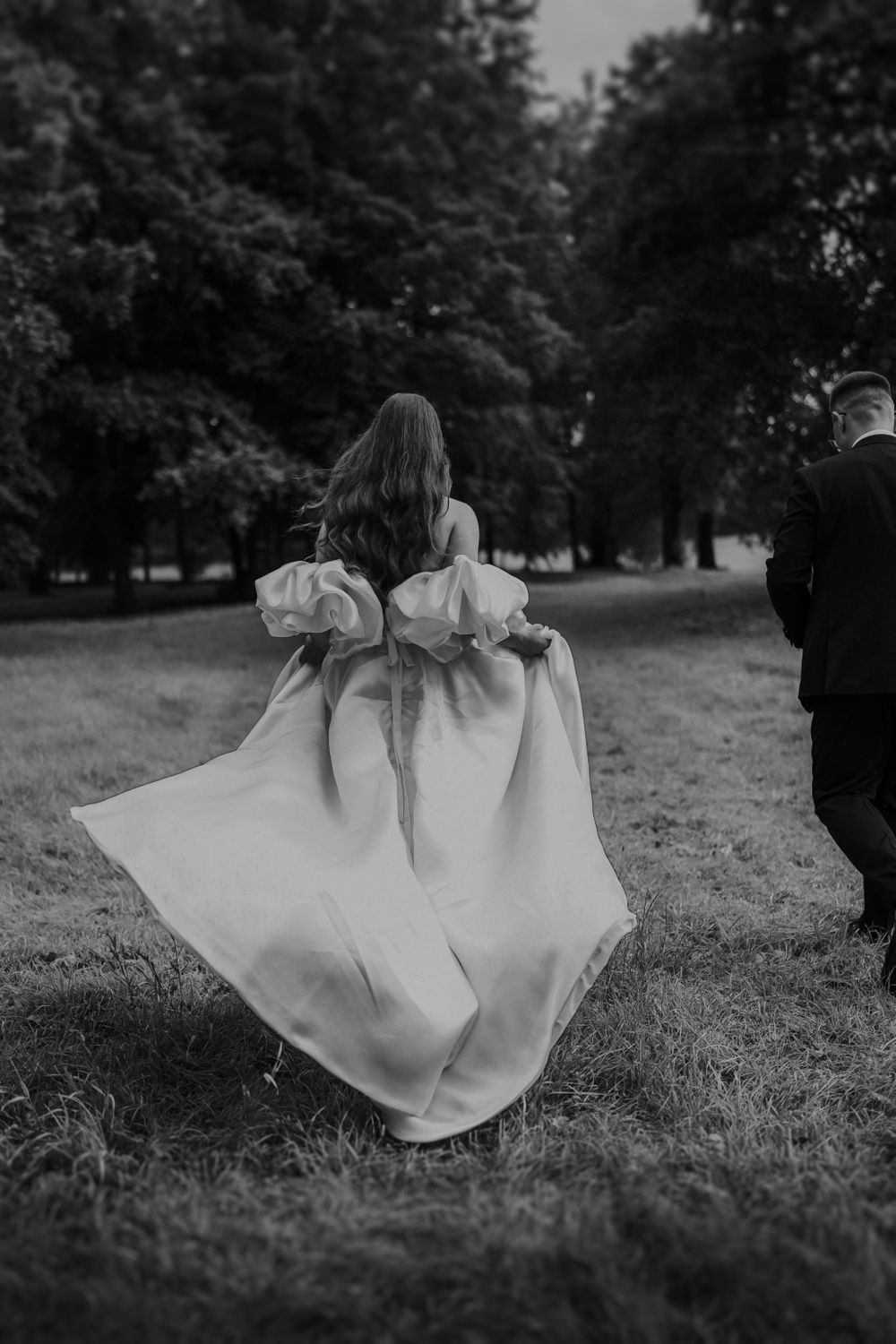 Bride's dress flows gracefully as couple walks through park.