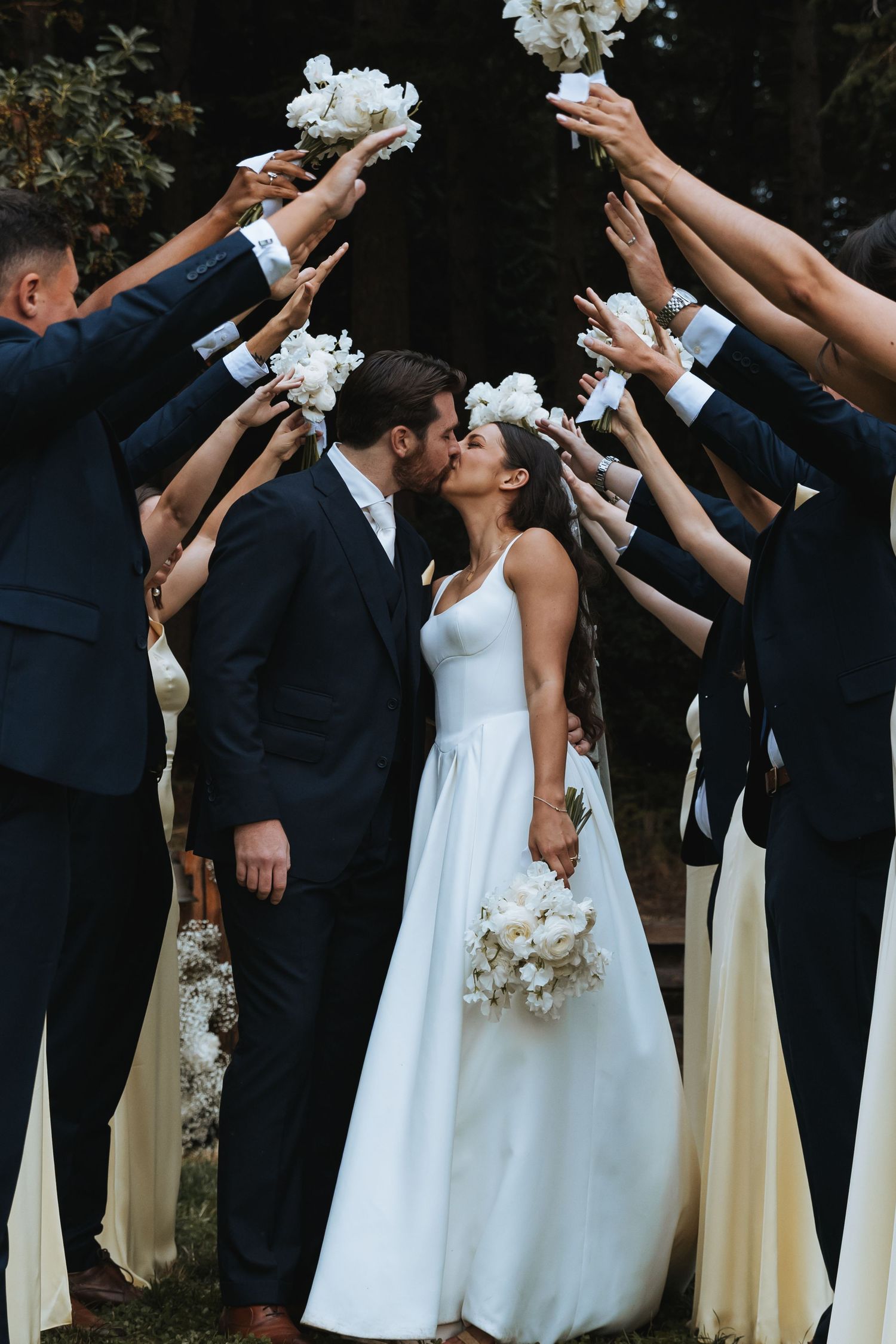 A bride and groom kiss under an arch of raised white flower bouquets held by their wedding party.