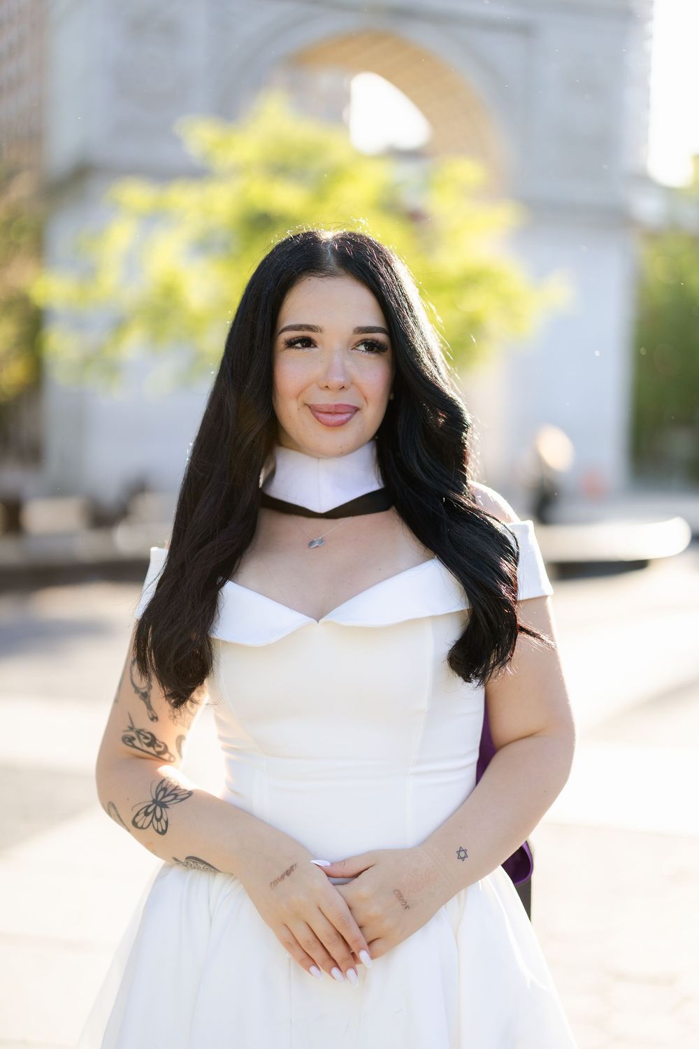 A person in a white dress with sheer neckline detail poses outdoors on a sunny day with blurred architecture in background.