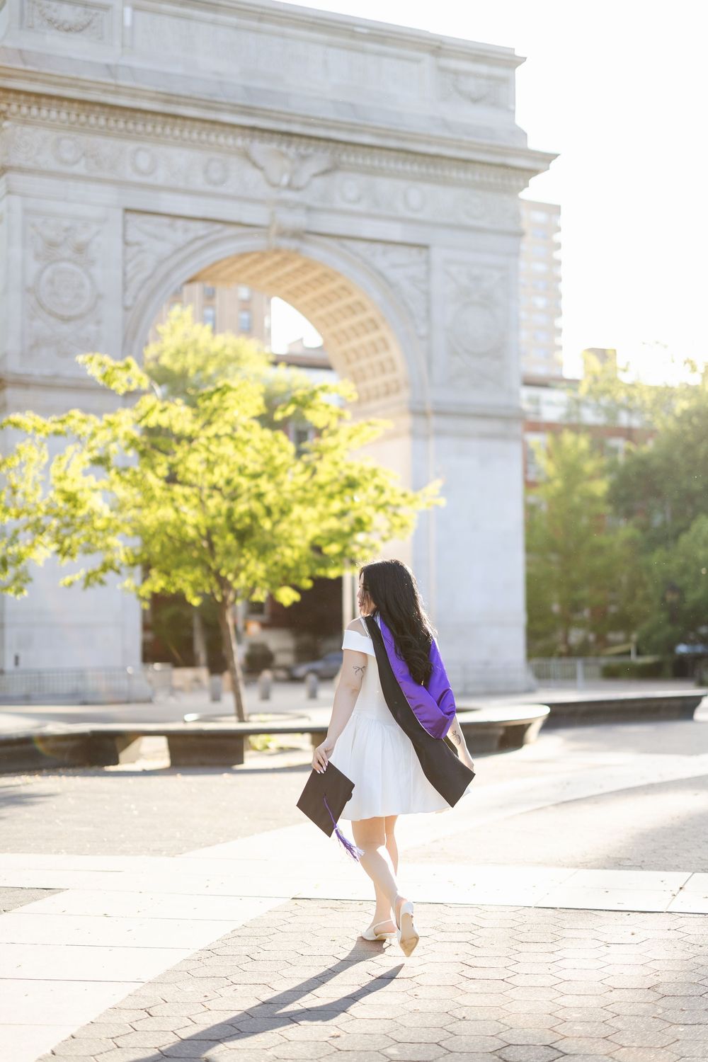 A graduate in cap and gown twirls joyfully in front of Washington Square Arch on a sunny spring day.