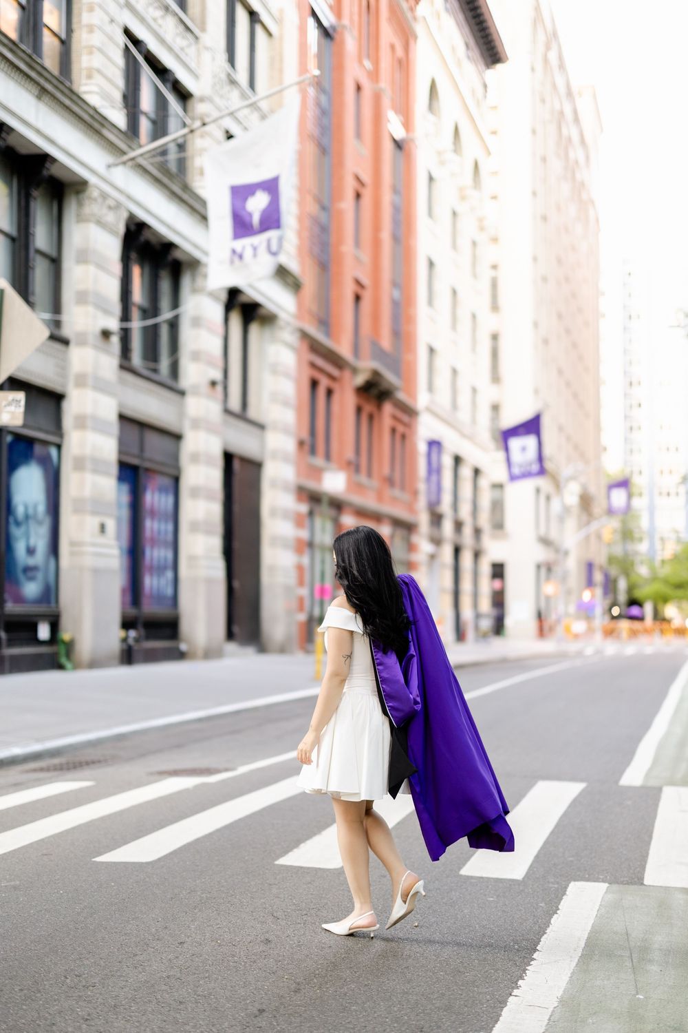A person in a white dress and purple graduation gown walks across a city street with brick buildings in the background.
