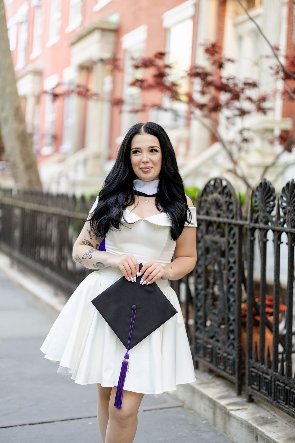 A woman in a white dress poses with graduation cap against an ornate iron fence on a city street.
