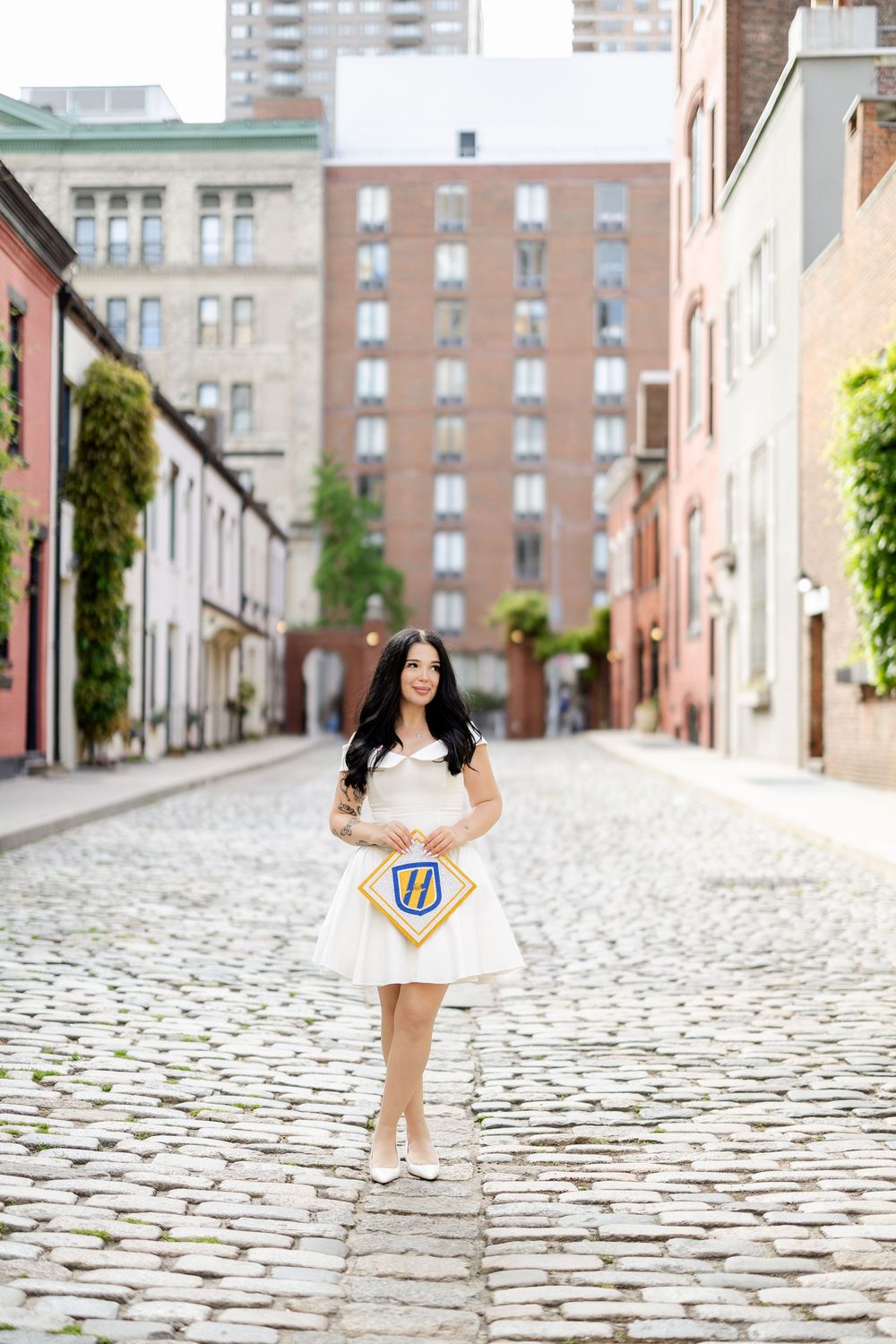 Graduate in white dress stands on cobblestone street with urban buildings in background.