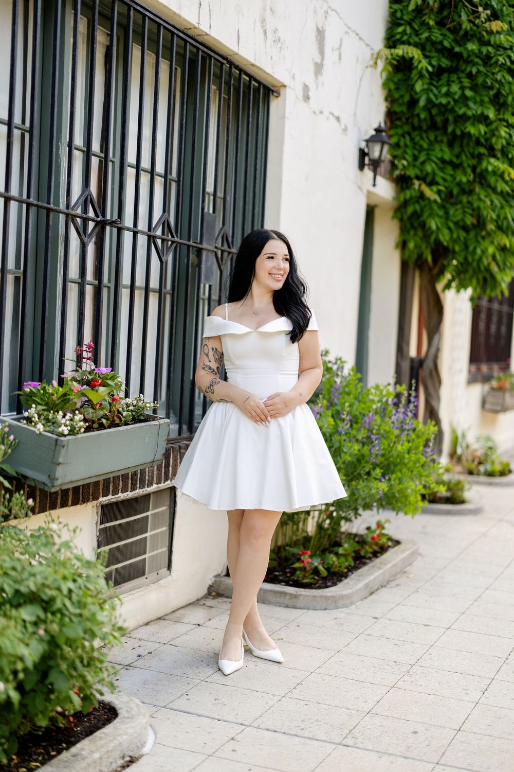 Woman in white summer dress poses along urban sidewalk with greenery and iron window bars in the background.