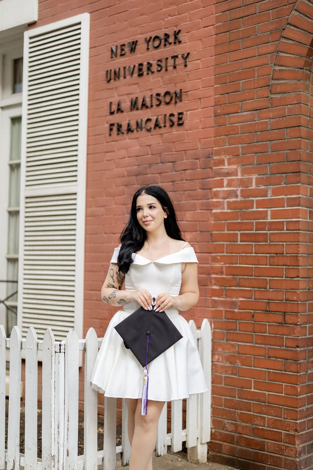 A graduate in a white dress poses with a graduation cap in front of NYU La Maison Francaise brick building.